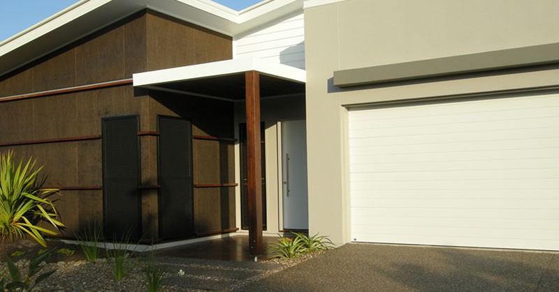 Modern house exterior with a brown and white facade, flat roof, a white garage door, and a small landscaped area with plants.