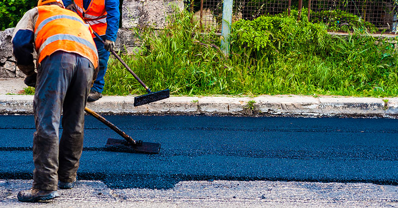 Two workers in safety vests use tools to spread hot asphalt while resurfacing a road next to a sidewalk and greenery.