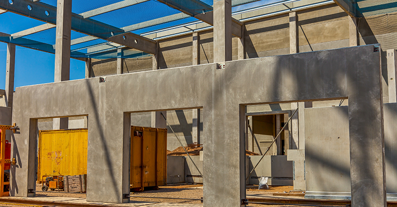 Partially constructed concrete building with exposed beams, three window or door openings, and a yellow container inside the structure under a blue sky.