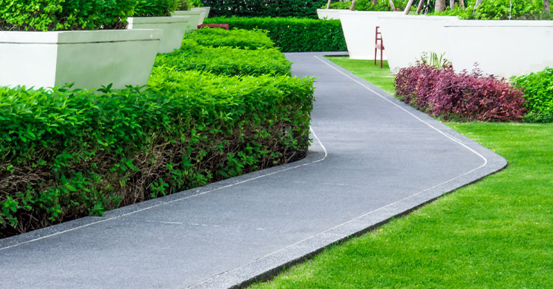 A paved walkway curves through a landscaped garden with trimmed green hedges, grass, and large rectangular planters.